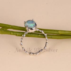 K&ouml;nnte beinhalten: Silberring mit einem ovalen Opal-Edelstein, der blaue und gr&uuml;ne Farbt&ouml;ne aufweist. Der Ring hat ein Band mit einem strukturierten, perlenbesetzten Design. Der Text "Anshik 925 Silver" ist sichtbar.