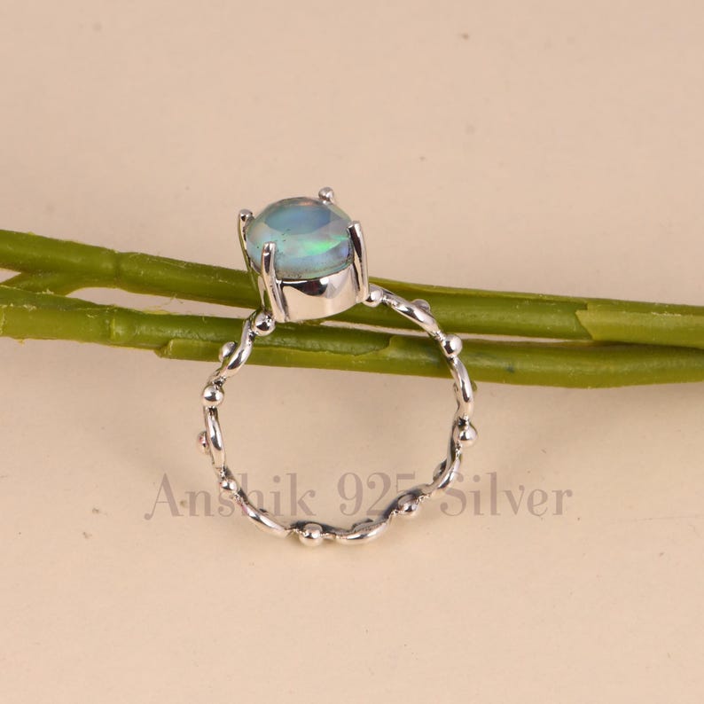 K&ouml;nnte beinhalten: Silberring mit einem ovalen Opal-Edelstein, der blaue und gr&uuml;ne Farbt&ouml;ne aufweist. Der Ring hat ein Band mit einem strukturierten, perlenbesetzten Design. Der Text "Anshik 925 Silver" ist sichtbar.