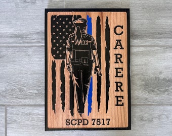 Puede incluir: Una placa de madera con una silueta negra de un policía de pie frente a una bandera estadounidense con una línea azul. El texto "CARRERE" está en el lado derecho de la placa y "SCPD 7517" está en la parte inferior.