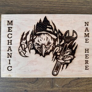 May include: A light-coloured wooden sign with a laser-cut design. It features an eagle with an American flag, holding a wrench. The word "MECHANIC" is vertically on the left, and "NAME HERE" is on the right. The sign is made of light-coloured wood.