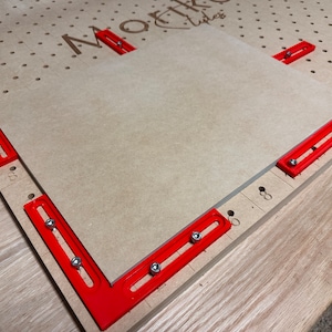 May include: A red woodworking jig with four clamps holding a piece of wood in place on a pegboard surface. The pegboard has a logo that says "MoFtK" and "Tiles".