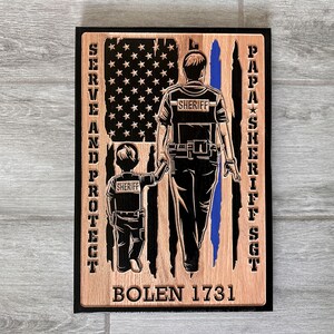 May include: Wooden sign with an American flag design featuring a sheriff and a child walking together. The text "SERVE AND PROTECT" is on the left side, "PAPA SHERIFF SGT" is on the right side, and "BOLEN 1731" is at the bottom.