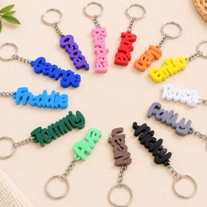 May include: A collection of twelve colorful, three-dimensional name keychains arranged in a circle. Each keychain features a custom name in a bold, rounded font attached to a silver-toned metal split ring by a short chain link.