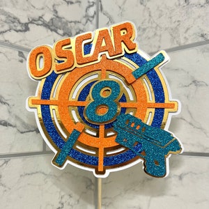 Könnte beinhalten: Ein Tortendekor mit einer Zielscheibe in Blau und Orange, der Zahl 8 in der Mitte und dem Namen "OSCAR" oben. Rechts neben der Zielscheibe befindet sich eine blaue Glitzer-Spielzeugpistole.