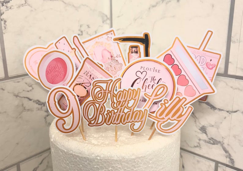 May include: Pink and gold cake toppers with the words "Happy Birthday Lilly" and various makeup and beauty themed designs.