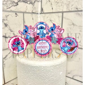 Puede incluir: Un pastel blanco con adornos de cupcake de tem&aacute;tica rosa y azul con Stitch y Angel de la pel&iacute;cula de Disney Lilo & Stitch. Los adornos tienen un dise&ntilde;o floral rosa y azul y dicen "Happy Birthday Lacey".