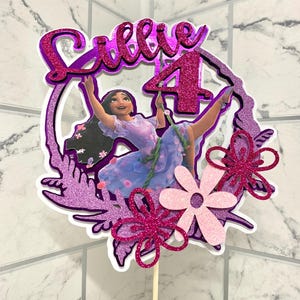 May include: Purple and pink cake topper featuring a cartoon image of a girl with long brown hair, a purple dress, and a pink flower in her hair. The topper also includes the name "Ellie" and the number "4" in glitter.