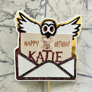 Könnte beinhalten: Eine weiße Eule mit goldenen Flügeln trägt einen roten und weißen Umschlag mit dem Text "Happy Birthday Katie" und dem Hogwarts-Wappen.