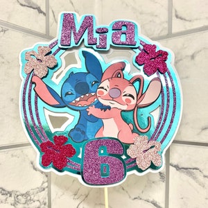 Puede incluir: Un adorno para pastel azul y rosa con Stitch y Angel, un 6 de purpurina morada y el nombre Mia en purpurina morada. El adorno est&aacute; sobre un fondo azul con flores rosas y blancas.