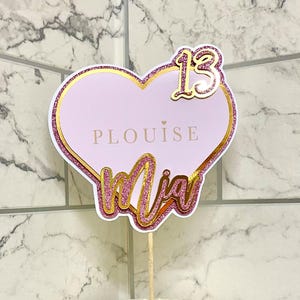May include: Pink and gold heart-shaped cake topper with the name "Mia" and the number "13" on it. The topper also has the text "PLOUISE" on it.