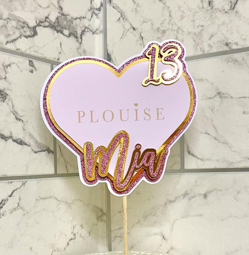 May include: Pink and gold heart-shaped cake topper with the name "Mia" and the number "13" on it. The topper also has the text "PLOUISE" on it.