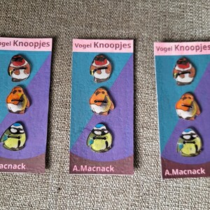 Op de afbeelding: Drie kaarten met vogelvormige knopen. Elke kaart heeft drie knopen: een rood-witte vogel, een oranje vogel en een blauw-gele vogel. De kaarten dragen de tekst "Vogel Knoopjes" en "A.Macnack".