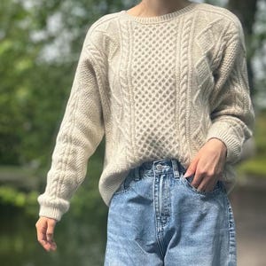 Peut inclure: Un pull en tricot de couleur crème avec un motif en losanges sur le devant. Le pull a des manches longues et un col rond. La personne porte un jean en denim bleu clair.