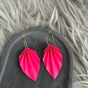 Könnte beinhalten: Ein Paar rosa Origami-Blatt-Ohrringe mit goldenen Kreisen. Die Ohrringe liegen auf einem schwarzen Tablett.
