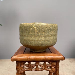 Pode incluir: Uma tigela de cerâmica texturizada, em tons de bege e verde. Repousa sobre um pequeno suporte de madeira finamente esculpido. As pernas do suporte têm um design semelhante ao bambu. A aparência rústica da tigela é realçada.