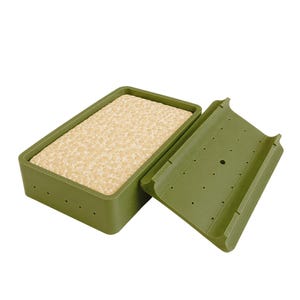 Puede incluir: Un molde rectangular de plástico verde oliva con su tapa a juego colocada al lado. El recipiente contiene un bloque de tempeh beige, poroso y texturizado. Tanto el recipiente como la tapa presentan pequeños orificios de ventilación.