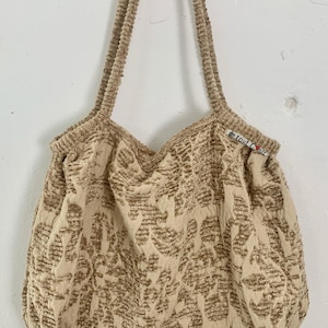 Op de afbeelding: Beige draagtas met corduroy bandjes en een textuurpatroon. De tas heeft een afgeronde vorm en een label met de tekst "de tout coeur". De tas hangt aan een houten plank met metalen haken.