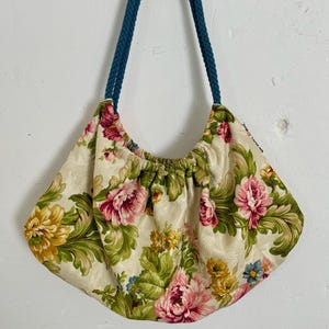 Może przedstawiać: Torebka w kwiatowy wzór z kremowym tłem i różowymi, żółtymi i zielonymi kwiatami. Torba ma marszczony top i niebieskie sznurkowe uchwyty. Torba wisi na drewnianym wieszaku z mosiężnymi haczykami.