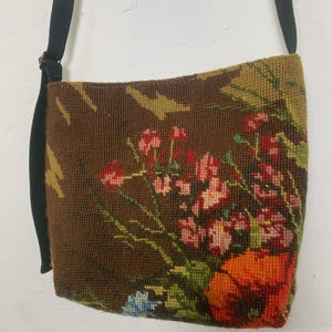 Puede incluir: Bolso de hombro de tapiz marrón con una correa negra. El bolso presenta un diseño floral con flores rojas, rosas, naranjas y azules. El fondo es una mezcla de marrón y verde.