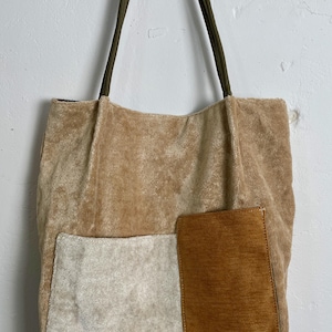 Może przedstawiać: Beżowa torba typu tote o miękkim, teksturowanym wyglądzie. Posiada dwie prostokątne naszywki w kontrastujących kolorach: złamanej bieli i brązu. Torba ma oliwkowozielone uchwyty.