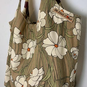 Op de afbeelding: Een tote bag met een bloemenpatroon. De tas heeft een bruine achtergrond met witte bloemen en groene stelen. De tas heeft beige bandjes en een klein label met de tekst "de tout cœur".