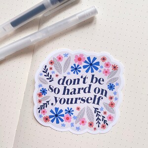 Puede incluir: Una pegatina blanca con un borde floral azul y el texto "no seas tan duro contigo mismo" en azul marino.