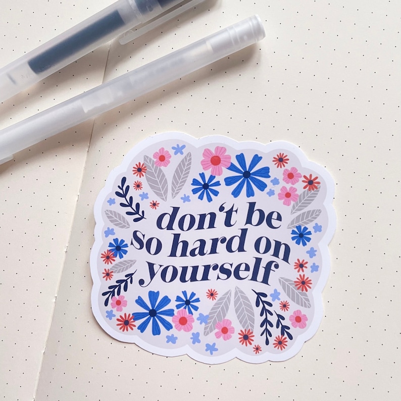 Puede incluir: Una pegatina blanca con un borde floral azul y el texto "no seas tan duro contigo mismo" en azul marino.