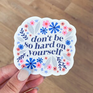 Puede incluir: Una pegatina blanca con un dise&ntilde;o floral azul y rosa. La pegatina dice "no seas tan duro contigo mismo".