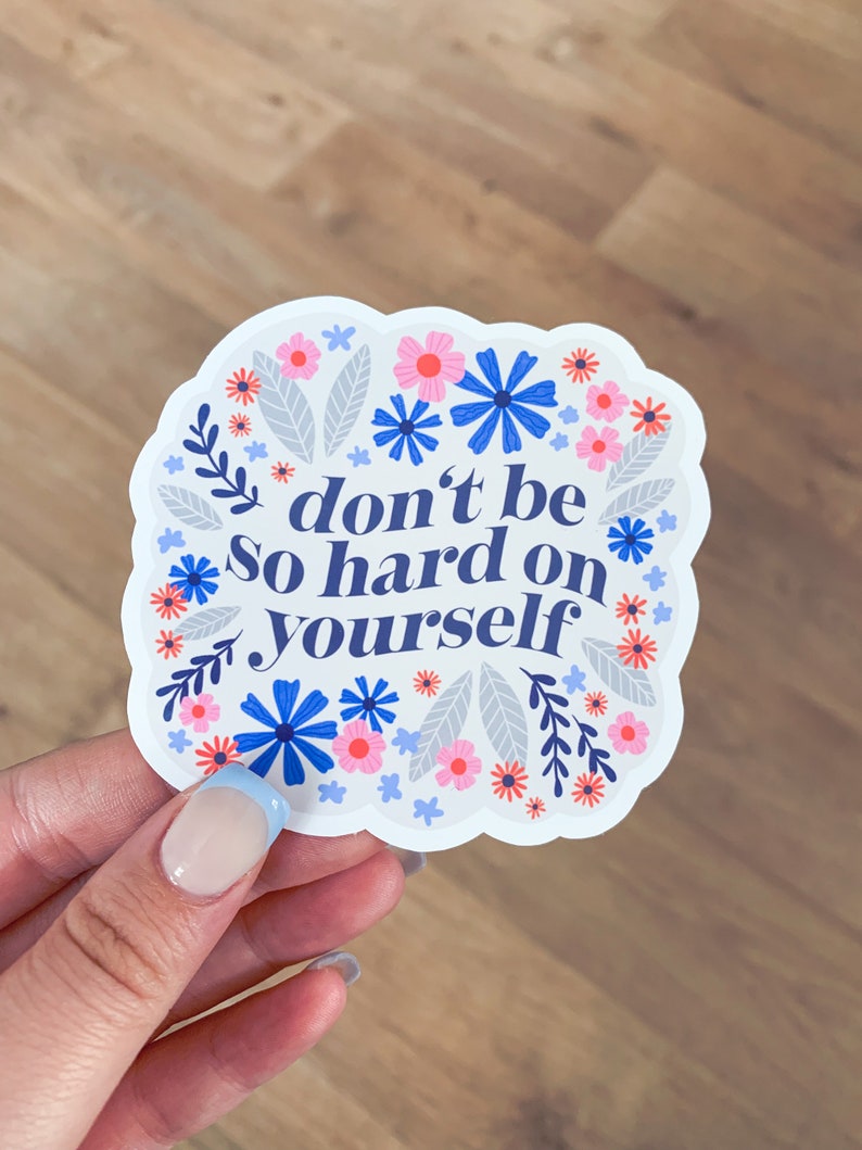 Puede incluir: Una pegatina blanca con un dise&ntilde;o floral azul y rosa. La pegatina dice "no seas tan duro contigo mismo".