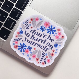 Puede incluir: Una pegatina con un dise&ntilde;o floral y el texto "no seas tan duro contigo mismo".