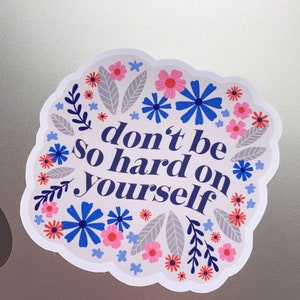 Puede incluir: Una pegatina blanca con un borde floral azul y el texto "no seas tan duro contigo mismo" en azul.
