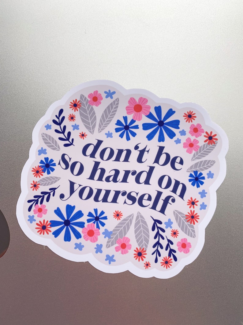 Puede incluir: Una pegatina blanca con un borde floral azul y el texto "no seas tan duro contigo mismo" en azul.