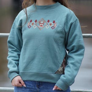 Puede incluir: Sudadera de color turquesa con un diseño geométrico bordado en rojo, blanco y marrón. El diseño está centrado en el pecho. La sudadera tiene cuello redondo y puños acanalados. Un panel de tela de camuflaje está cosido en la costura lateral.