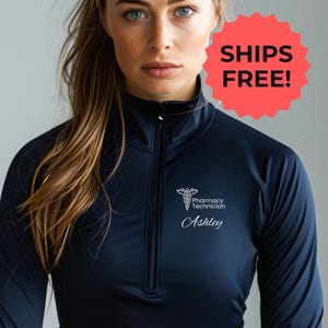 Peut inclure: Haut de sport bleu marine avec fermeture éclair quart de zip. Il présente un symbole de pharmacie blanc, les mots "Pharmacy Technician" et le nom "Ashley". Col montant et fermeture éclair sur la poitrine.