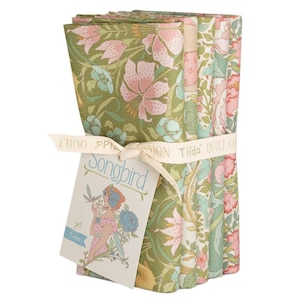 Könnte beinhalten: Ein Stapel Stoffquadrate mit Blumenmuster, gebunden mit einem cremefarbenen Band. Der Stoff zeigt ein Muster aus rosa Lilien und blauen Blumen auf grünem Hintergrund. Ein Etikett mit dem Wort "Songbird" und einer Frauenillustration ist angebracht.