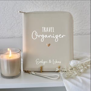 Könnte beinhalten: Ein cremefarbener Reiseorganizer mit Reißverschluss. Auf der Vorderseite des Organizers steht der Text "TRAVEL Organizer" in weißer Kursivschrift mit einem kleinen goldenen Herzen. Unter dem Text steht der Name "Evelyn & Lukas" in weißer Schrift.