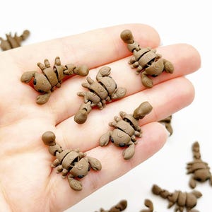 May include: Brown scorpion-shaped polymer clay charms. The charms are detailed with segmented bodies, claws, and curved tails. Several charms are held in a hand, with others scattered on a white surface. These charms are suitable for jewelry making or craft projects.