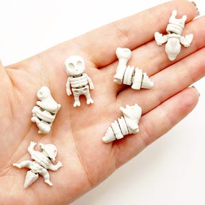 Op de afbeelding: Een hand houdt verschillende kleine, gebroken witte skeletfiguren vast. De figuren omvatten een menselijk skelet, een dinosaurus en andere wezens. De figuren zijn gemaakt van een hard materiaal en hebben gedetailleerde skeletkenmerken.
