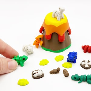 May include: A brown volcano model with yellow and orange lava, surrounded by small dinosaur figures in green, orange, blue, and red, along with white bone-shaped pieces, brown fossil discs, and small yellow leaf-shaped accents on a white surface.