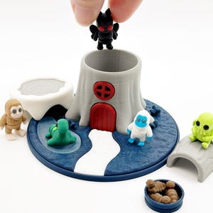 May include: A miniature fantasy playset featuring a gray tree stump with a red door, surrounded by small figurines of various creatures. The base is blue with a white path. A hand is placing a black figure.