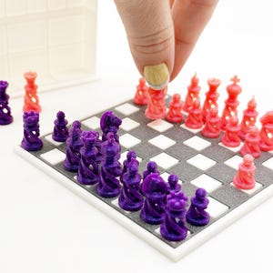 Könnte beinhalten: Miniatur-Schachspiel mit lila und roten Figuren auf einem karierten Brett. Die Figuren sind auf dem Brett angeordnet, wobei eine Hand eine rote Figur platziert. Eine weiße Aufbewahrungsbox befindet sich im Hintergrund.