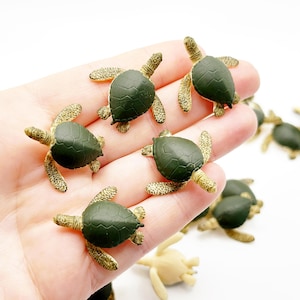 May include: Miniature sea turtles with dark green shells and tan flippers are displayed. The small turtle figurines are arranged in a hand, with several more visible in the background. The turtles are a charming decorative item.