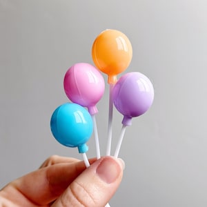 May include: Four colorful balloon cake toppers, in blue, pink, purple, and orange, on white sticks. The balloons are glossy and have a slight shine.