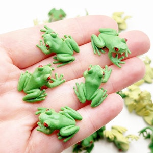 May include: A hand holding a collection of small green plastic frog figurines with red eyes. The frogs are arranged in a variety of poses.