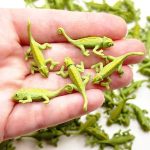 Könnte beinhalten: Eine Hand hält eine Sammlung kleiner grüner Plastik-Chamäleons. Die Chamäleons haben gelbe Streifen und sind etwa 5 cm lang.