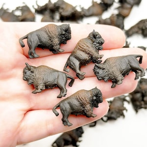 May include: A collection of small, detailed bison figurines. The miniature sculptures are primarily dark brown with lighter brown fur detailing on their heads and shoulders. The bison are in various poses, suggesting movement.