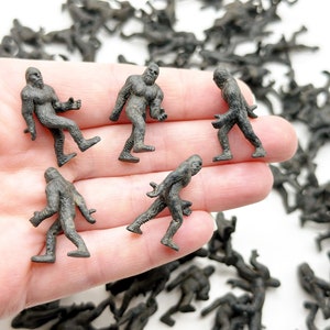 May include: A hand holding five small, gray plastic figurines of Bigfoot. The figurines are all in different poses and are about 2 inches tall.