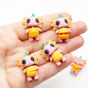 Peut inclure: Plusieurs petites figurines d'axolotl colorées. Chaque figurine a une tête rose avec des yeux noirs, un corps jaune et des franges orange. Les axolotls sont dans diverses poses, avec une tige verte sur le dessus.