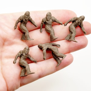 May include: Six brown miniature figurines of Bigfoot, each standing upright with arms outstretched, on a person's hand.