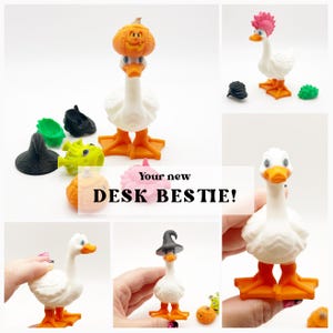 May include: A white goose figurine with orange feet and beak, featuring interchangeable accessories including a pumpkin, a witch hat, a pink mohawk, and a green spiky piece. The text "Your new DESK BESTIE!" is displayed over the collection.
